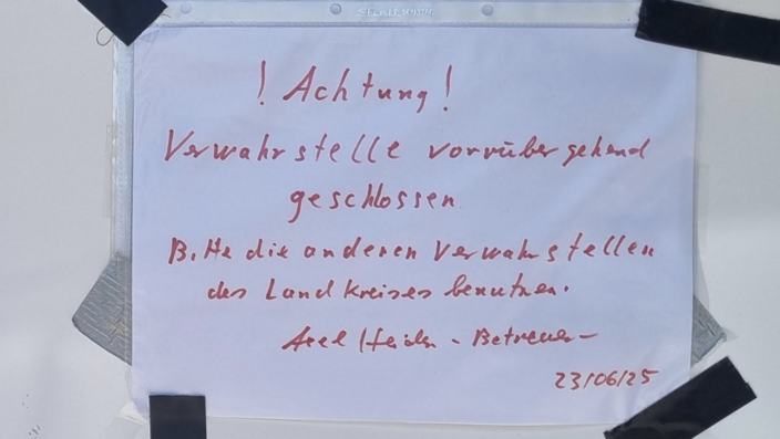 Ein Zettel, handgeschrieben, informiert über die vorübergehende Schließung einer Verwahrstelle. Er rät zur Nutzung anderer Stellen im Landkreis, datiert auf 23/06/25. Text oben: "Achtung!"