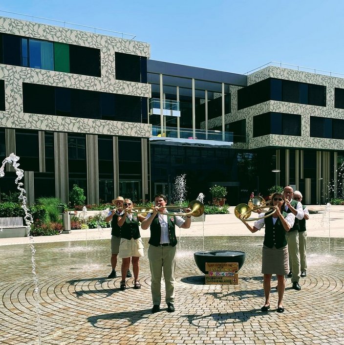 Eine Blaskapelle spielt Musik im Freien vor einem modernen Gebäude mit großen Fenstern. Im Vordergrund sprühen kleine Springbrunnen Wasser. Ein Schild zeigt „Pfullendorf Stadtorchester“.